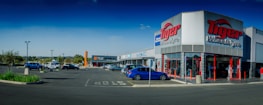 A commercial shopping area with a 'Tiger Wheel & Tyre' store prominently in the foreground, featuring large signage. Several other storefronts are visible in the background, including 'Tile Africa'. The parking lot has a few parked cars, and the weather appears clear with a deep blue sky.