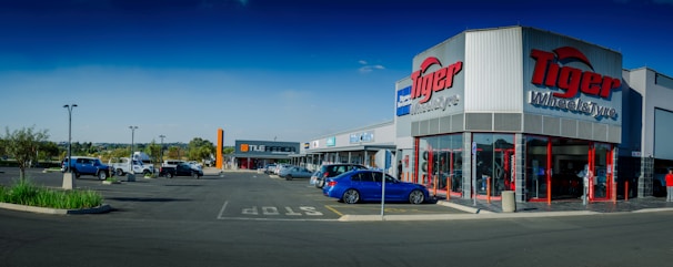A commercial shopping area with a 'Tiger Wheel & Tyre' store prominently in the foreground, featuring large signage. Several other storefronts are visible in the background, including 'Tile Africa'. The parking lot has a few parked cars, and the weather appears clear with a deep blue sky.