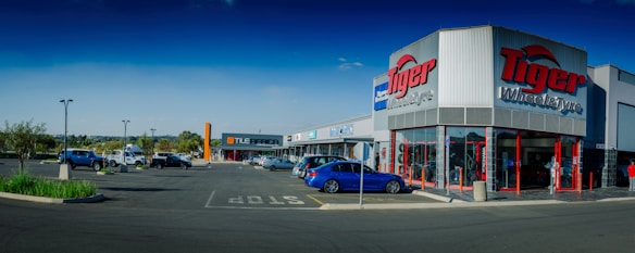 A commercial shopping area with a 'Tiger Wheel & Tyre' store prominently in the foreground, featuring large signage. Several other storefronts are visible in the background, including 'Tile Africa'. The parking lot has a few parked cars, and the weather appears clear with a deep blue sky.