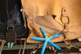 A collection of items including a textured brown leather bag with metal rings, a vibrant blue starfish lying on top, various black leather and chain accessories, and a pair of triangular turquoise earrings.