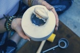 A person holding a freshly opened coconut with a piece of coconut flesh partially removed, revealing the water inside. The individual is wearing bracelets made of different colored beads and has manicured nails. Nearby, there is a metal tool, likely used to open the coconut.