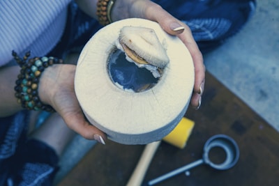 A person holding a freshly opened coconut with a piece of coconut flesh partially removed, revealing the water inside. The individual is wearing bracelets made of different colored beads and has manicured nails. Nearby, there is a metal tool, likely used to open the coconut.