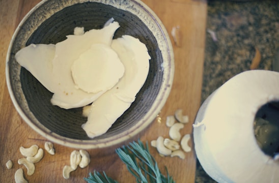 A bowl containing pieces of white coconut meat is placed on a wooden surface. Surrounding the bowl are cashew nuts and a sprig of rosemary. To the side, a partially opened young coconut is visible.