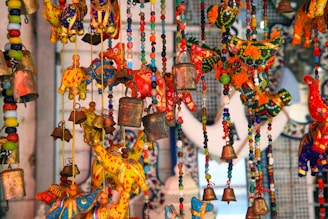 An array of colorful handcrafted decorative items, featuring vibrant fabric elephants and birds, intricately designed with beads and small metallic bells. The scene is lively with a mix of reds, yellows, blues, and greens creating a vibrant tapestry.