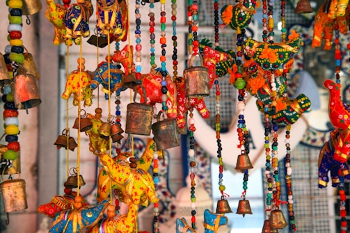 An array of colorful handcrafted decorative items, featuring vibrant fabric elephants and birds, intricately designed with beads and small metallic bells. The scene is lively with a mix of reds, yellows, blues, and greens creating a vibrant tapestry.