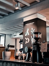 A modern café interior with a coffee grinder and brewing equipment on the countertop. The wall features illustrations related to the Heimlich maneuver and a plant in a pot adds a touch of greenery. Exposed beams and industrial elements contribute to a contemporary aesthetic.