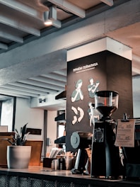 A modern café interior with a coffee grinder and brewing equipment on the countertop. The wall features illustrations related to the Heimlich maneuver and a plant in a pot adds a touch of greenery. Exposed beams and industrial elements contribute to a contemporary aesthetic.