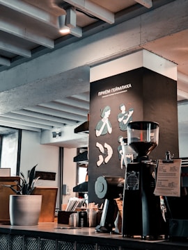 A modern café interior with a coffee grinder and brewing equipment on the countertop. The wall features illustrations related to the Heimlich maneuver and a plant in a pot adds a touch of greenery. Exposed beams and industrial elements contribute to a contemporary aesthetic.
