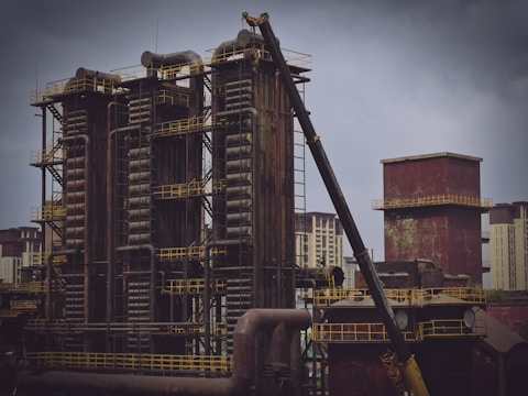 A large industrial facility with several tall, rusted metal structures and chimneys surrounded by a complex network of pipes and ladders. A crane is positioned in the foreground, suggesting construction or maintenance work. The sky is overcast, adding a gloomy atmosphere.