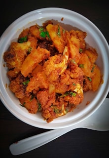 A white bowl filled with a spicy and seasoned dish consisting of golden-brown fried cauliflower and potatoes, garnished with green herbs. The bowl is accompanied by a white spoon placed on a dark surface.