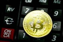 A golden Bitcoin coin is placed on a black calculator. The calculator keys are visible, including a red ON/CA button and various numbers and symbols, such as a Euro symbol.