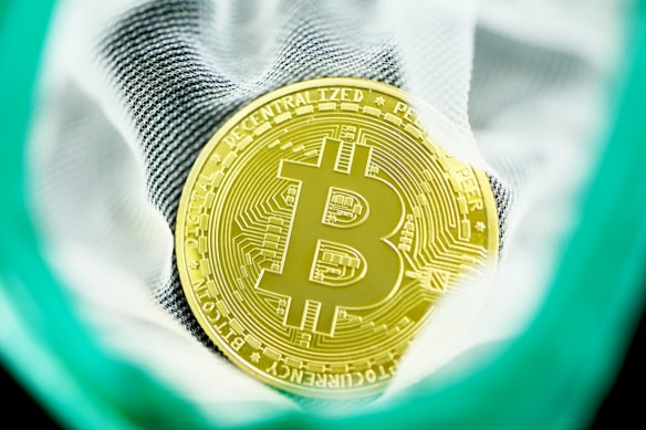A gold-colored Bitcoin coin with a prominent Bitcoin logo is surrounded by fabric material, possibly a bag. The coin features intricate circuit-like designs and is highlighted as the central focus of the image. The background shows a mesh pattern in close-up.