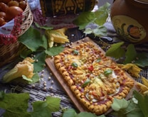 A rectangular pizza with vibrant toppings such as squash blossoms, cherry tomatoes, chickpeas, and fresh basil leaves sits on a wooden board. It is surrounded by green leaves and scattered chickpeas on a patterned cloth. A basket filled with tomatoes is partially covered with a decorative cloth, and a painted ceramic pot is visible in the background.