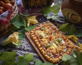 A rectangular pizza with vibrant toppings such as squash blossoms, cherry tomatoes, chickpeas, and fresh basil leaves sits on a wooden board. It is surrounded by green leaves and scattered chickpeas on a patterned cloth. A basket filled with tomatoes is partially covered with a decorative cloth, and a painted ceramic pot is visible in the background.