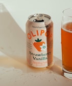 A can of Olipop Strawberry Vanilla sparkling tonic sits next to a glass partially filled with the drink. The can features colorful illustrations including a strawberry and vanilla flower, and highlights health-related benefits such as prebiotics and botanicals.