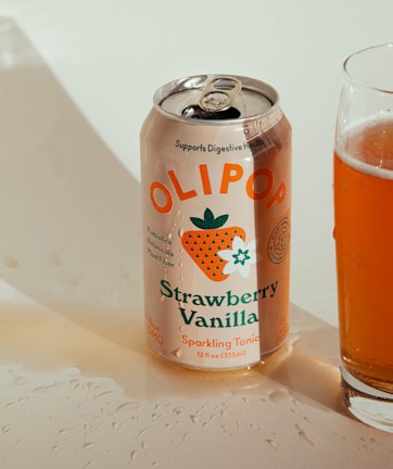 A can of Olipop Strawberry Vanilla sparkling tonic sits next to a glass partially filled with the drink. The can features colorful illustrations including a strawberry and vanilla flower, and highlights health-related benefits such as prebiotics and botanicals.