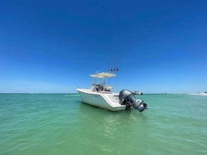 A motorboat with several fishing rods is anchored in clear, turquoise water under a cloudless blue sky. The boat's outboard motor is visible, and there are other distant boats on the horizon.
