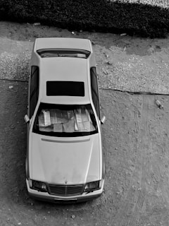 A black and white image of a car parked on a dirt surface, viewed from above. The windshield is covered with papers or reflective material. The surroundings include a pathway and vegetation.