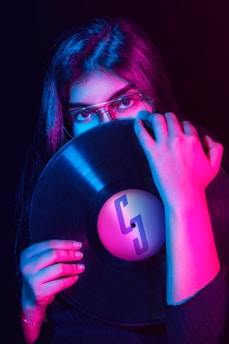 A person with long dark hair and glasses holds a vinyl record partially covering their face. The scene is bathed in vibrant pink and blue lighting, casting colorful reflections on the vinyl surface and the person's hands.