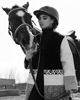 A person in equestrian attire is standing beside a horse. The individual is wearing a helmet and a patterned coat, holding the horse by the reins. The horse has a strikingly marked face with a white blaze. The image is in black and white, creating a classic and elegant feel.