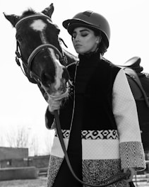 A person in equestrian attire is standing beside a horse. The individual is wearing a helmet and a patterned coat, holding the horse by the reins. The horse has a strikingly marked face with a white blaze. The image is in black and white, creating a classic and elegant feel.