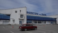 A seemingly abandoned or quiet airport terminal is pictured with a vintage red car parked in the forefront. The building is white with blue glass windows and displays a large sign reading 'ROSTOV ON DON' on the roof. The sky is overcast, and there are a few traffic cones placed around the parking area.