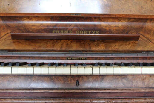 A wooden piano with a rich, textured finish displays a row of white and black keys. The name 'Franz Goetze' is engraved above the keys on a panel with a polished wood grain surface, suggesting an elegant and classical instrument.