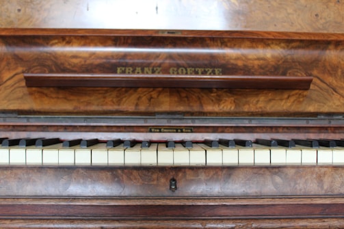 A wooden piano with a rich, textured finish displays a row of white and black keys. The name 'Franz Goetze' is engraved above the keys on a panel with a polished wood grain surface, suggesting an elegant and classical instrument.