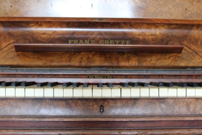 A wooden piano with a rich, textured finish displays a row of white and black keys. The name 'Franz Goetze' is engraved above the keys on a panel with a polished wood grain surface, suggesting an elegant and classical instrument.