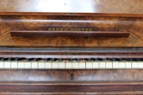 A wooden piano with a rich, textured finish displays a row of white and black keys. The name 'Franz Goetze' is engraved above the keys on a panel with a polished wood grain surface, suggesting an elegant and classical instrument.