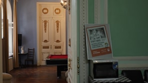 A room with ornate, classic decor featuring golden accents on white double doors. Inside, there is a billiards table with a red surface. A poster is propped up against an open door, displaying text. A small table holds a microwave, and a black chair is visible. Natural light enters through large windows with beige curtains.