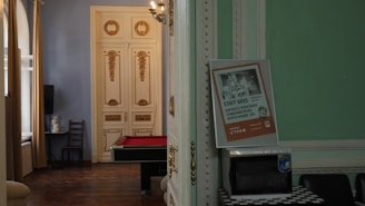 A room with ornate, classic decor featuring golden accents on white double doors. Inside, there is a billiards table with a red surface. A poster is propped up against an open door, displaying text. A small table holds a microwave, and a black chair is visible. Natural light enters through large windows with beige curtains.