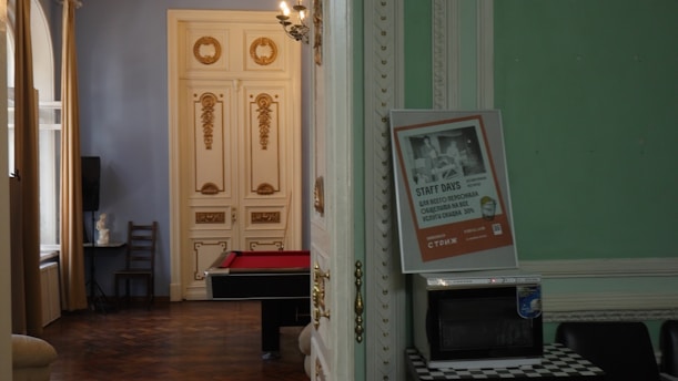 A room with ornate, classic decor featuring golden accents on white double doors. Inside, there is a billiards table with a red surface. A poster is propped up against an open door, displaying text. A small table holds a microwave, and a black chair is visible. Natural light enters through large windows with beige curtains.