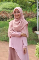 A woman wearing a light pink hijab and dress stands in a lush green garden. Her arms are crossed, and she is smiling softly, conveying a serene and peaceful presence.