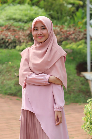 A cheerful young woman in a pastel pink hijab paired with a flowing floral midi skirt and a cozy knit sweater, standing in a sunlit park.