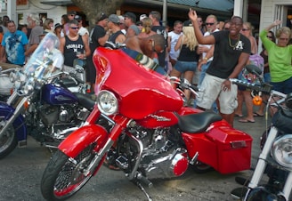 A crowded scene filled with people gathered around shiny motorcycles. The focal point is a bright red motorcycle with chrome accents. Individuals in casual attire appear to be enjoying an outdoor event, with some taking photos and others chatting.