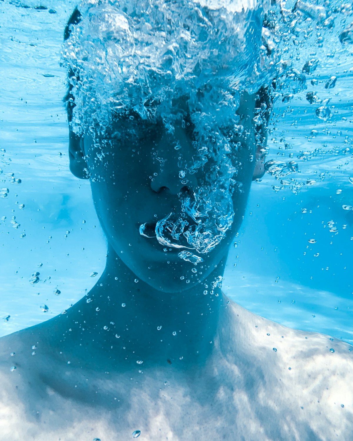 A person's face submerged underwater amid rising bubbles, bodily-transition register
