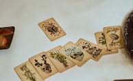 A set of intricately designed cards laid out on a light surface, each featuring illustrations of skulls intertwined with botanical elements. The cards are artistically detailed, showcasing a blend of anatomical and floral designs. To the left, there is a wooden plate with apple slices, and to the right, the edge of a dark ribbed circular dish is visible.