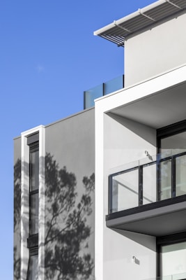 A modern architectural facade featuring clean lines and a minimalist design. The building has large glass windows and balconies, with the shadows of nearby trees cast on the grey walls. The sky is clear and bright blue, complementing the clean and contemporary aesthetic.