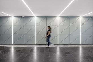 A modern hallway features a sleek design with a geometric pattern on the walls, divided by illuminated vertical lines. The floor is made of dark tiles, and a person is seen walking across, slightly blurred due to motion.