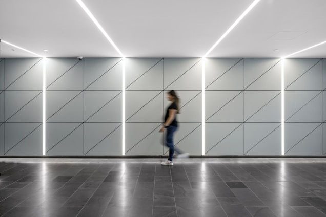 A modern hallway features a sleek design with a geometric pattern on the walls, divided by illuminated vertical lines. The floor is made of dark tiles, and a person is seen walking across, slightly blurred due to motion.