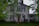 A two-story house with white siding and black shutters. The front door is wooden with a small set of steps leading up to it, flanked by small green bushes. An American flag is hung near the entrance. There is a tree in the front yard partially obscuring the view of the house. The lawn is green and well-maintained with other trees and shrubs around. A covered wooden porch is seen attached to the side of the house.