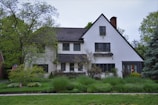 A two-story house with a steeply pitched roof and white stucco exterior surrounded by lush green vegetation. There are several trees and shrubs in the garden, creating a peaceful and natural setting. The house has multiple windows and a chimney, and an American flag is visible near the front.