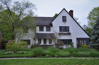 A two-story house with a steeply pitched roof and white stucco exterior surrounded by lush green vegetation. There are several trees and shrubs in the garden, creating a peaceful and natural setting. The house has multiple windows and a chimney, and an American flag is visible near the front.