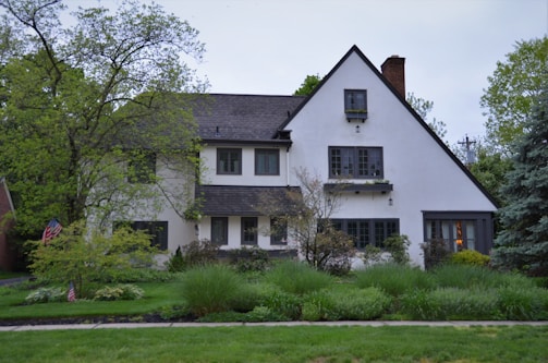 A two-story house with a steeply pitched roof and white stucco exterior surrounded by lush green vegetation. There are several trees and shrubs in the garden, creating a peaceful and natural setting. The house has multiple windows and a chimney, and an American flag is visible near the front.