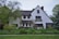A two-story house with a steeply pitched roof and white stucco exterior surrounded by lush green vegetation. There are several trees and shrubs in the garden, creating a peaceful and natural setting. The house has multiple windows and a chimney, and an American flag is visible near the front.