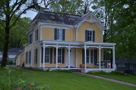 A charming, two-story Victorian-style house painted in a soft yellow hue with intricate white trim. The house features a covered porch with detailed woodwork and several large windows. It is surrounded by lush greenery with tall trees and a well-kept lawn.