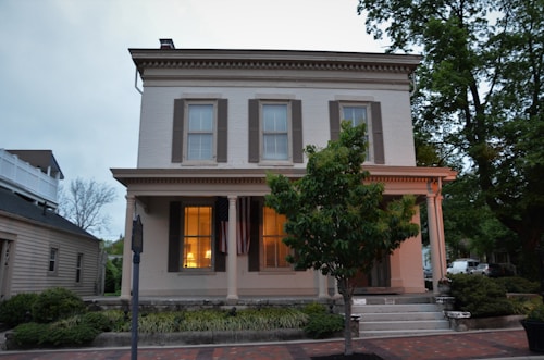 A two-story historic house with large windows and American flags hanging near the entrance. There is a well-maintained garden with shrubs and a tree in front of the house. The structure features a symmetrical design with white and beige colors, complemented by Greek Revival architectural elements.