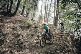 Two mountain bikers are navigating a rugged, wooded trail with a steep incline in a dense forest. They are wearing helmets and appropriate biking gear, demonstrating focus and skill as they maneuver around the terrain.
