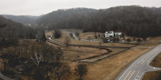 A rural landscape featuring a large two-story white house surrounded by expansive fields with sparse trees. A winding road cuts through the scene, leading past the house and into the distance. Hills densely covered with bare trees form the backdrop, under a gray, overcast sky.